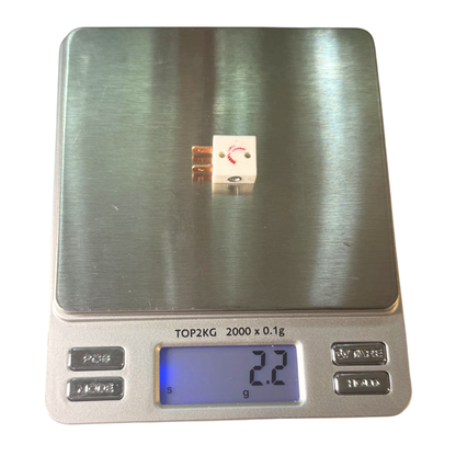 A small electronic power switch with copper-colored leads sits on the weighing platform of a digital scale. The scale has a brushed stainless steel top and a silver base with a blue-backlit LCD display showing 2.2g